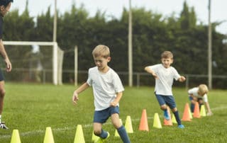 Mature male coach watching as pre-teen male footballers work on agility training with running drills around cones.