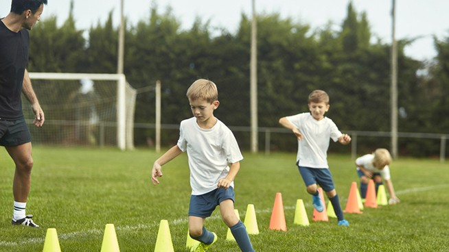 Mature male coach watching as pre-teen male footballers work on agility training with running drills around cones.