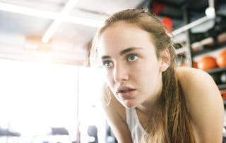 Beautiful young woman in gym, in white tank top, resting, sweat on her face.