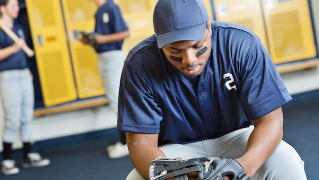 baseball player sitting in locker room looking distressed with teammates in background