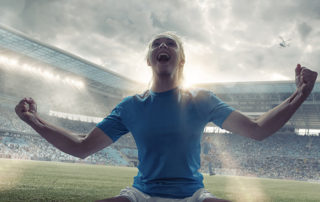 A close up cross processed image of a professional woman soccer player kneeling on the grass with her arms out, fists clenched and head back, shouting in happiness and celebration at winning. The player is in a generic floodlit stadium full of spectators under a cloudy evening sky.