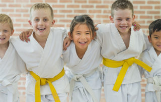 A group of elementary age children are taking a martial arts class. They are standing together in a row and are smiling while looking at the camera.