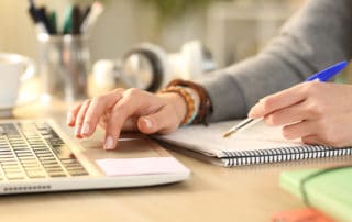 Close up of student girl hands comparing notes on notebook with laptop at home