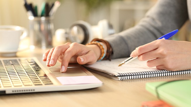 Close up of student girl hands comparing notes on notebook with laptop at home