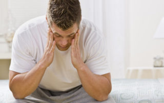 man sitting on edge of bed holding head with both hands due to lack of sleep